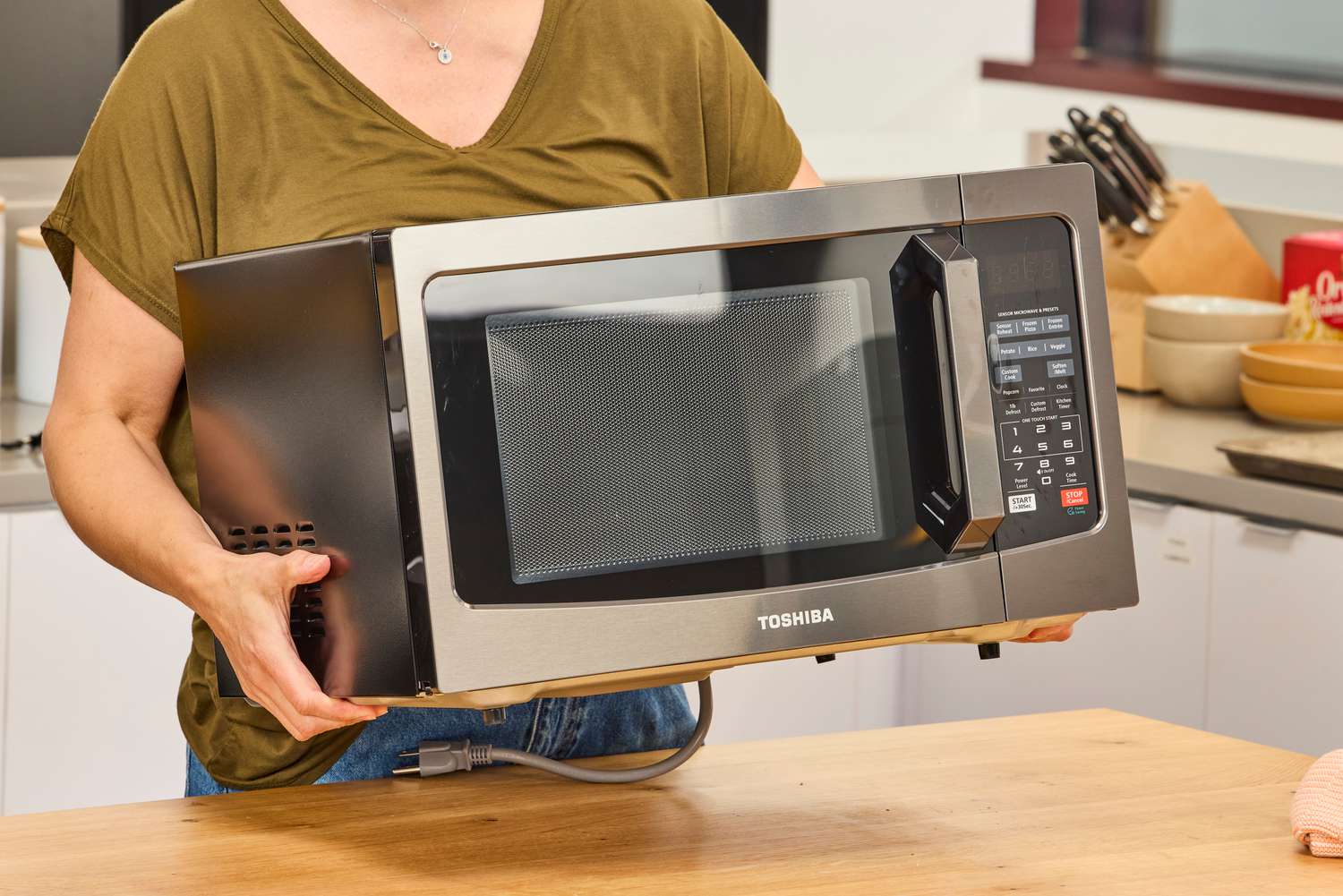 A person lifting the Toshiba EM131A5C-BS Microwave Oven above a wooden counter
