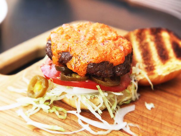 A fully loaded pimento burger with the top bun laying next to it. The burger has lettuce, tomato, and jalalpenos.