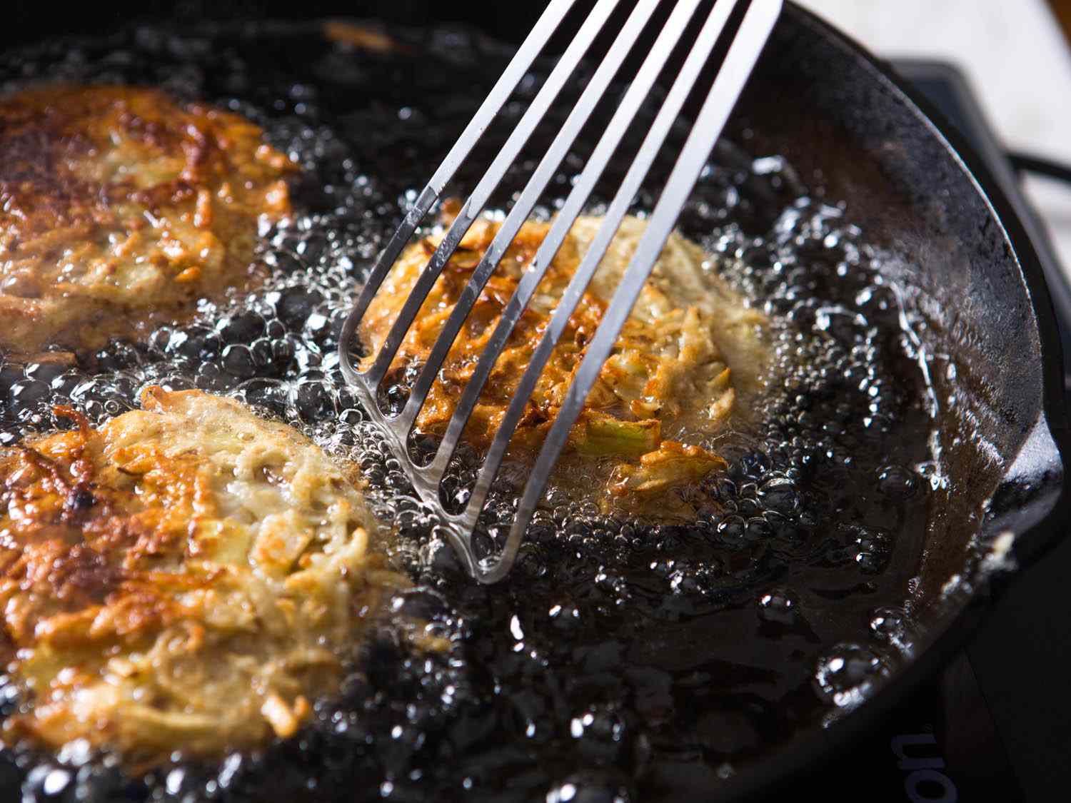 Three latkes browning and being moved in a cast iron skillet by a metal spatula. 