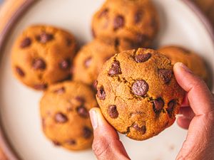 A closeup of hands holding a chocolate chip cookie with other cookies visible on a plate in the background