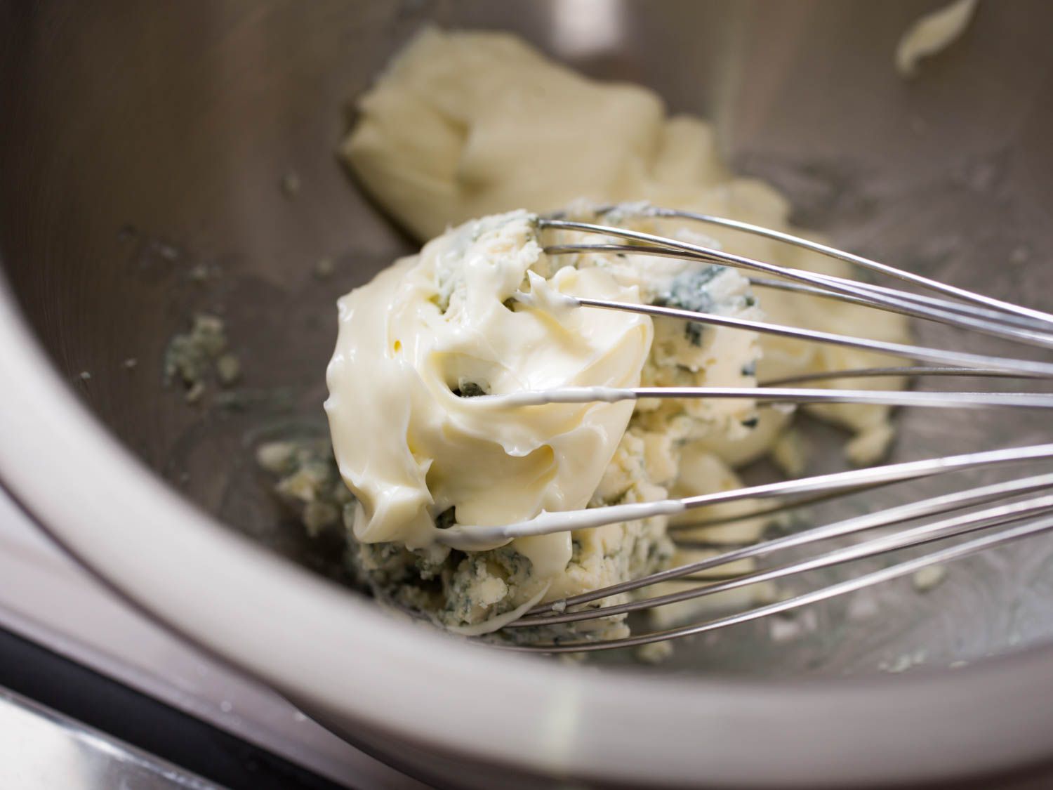 Mayonnaise, sour cream, and buttermilk being whisked with blue cheese in a metal bowl.