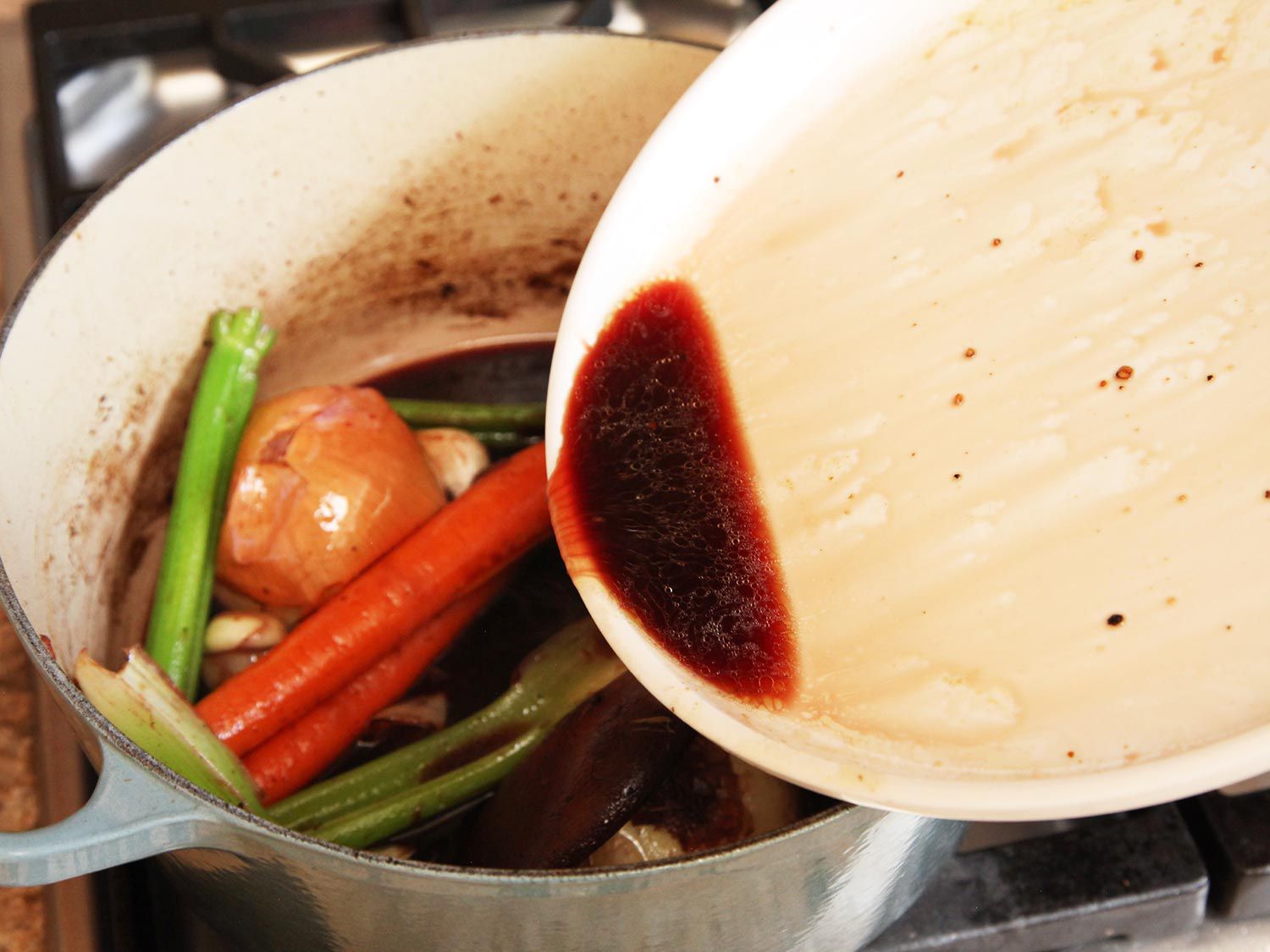 Pouring the juices from browned chuck roast from a white plate into a Dutch oven with vegetables for All-American beef stew.