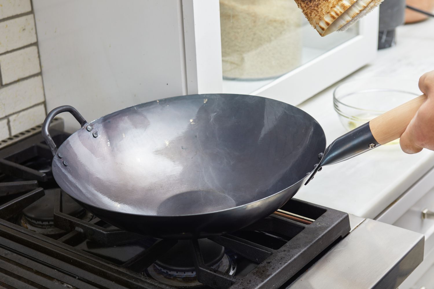 The Mammafong Pre-Seasoned Blue Carbon Steel Flat-Bottom Pow Wok displayed on a stove with a hand holding the handle