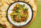 Bowl of pickle dip garnished with panko, dill and pickles, surrounded by a wooden plate full of potato chips, on a green tabeltop