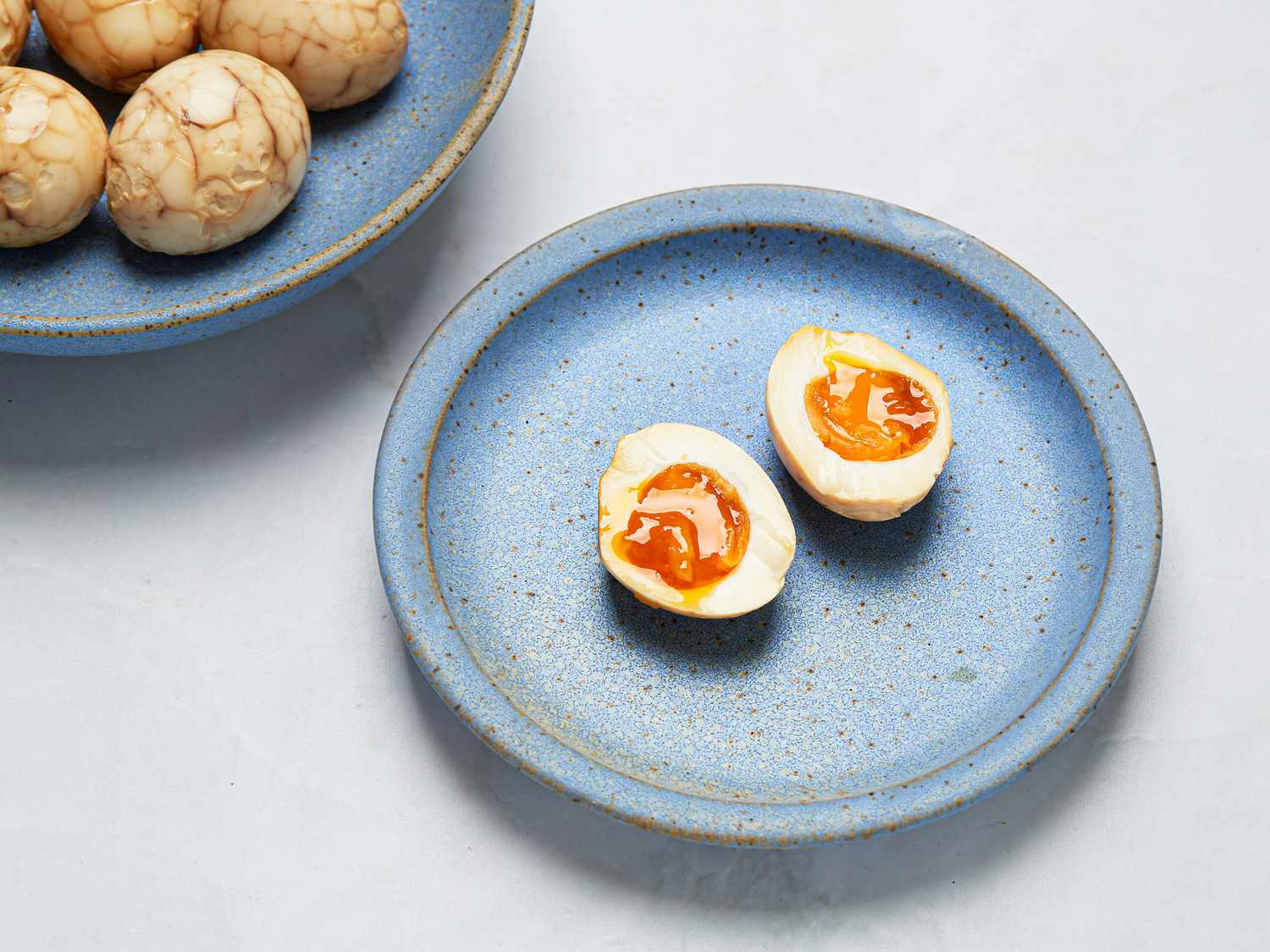 Blue plate of sliced open tea egg, with a blue bowl of whole eggs to the side, on a white surface 