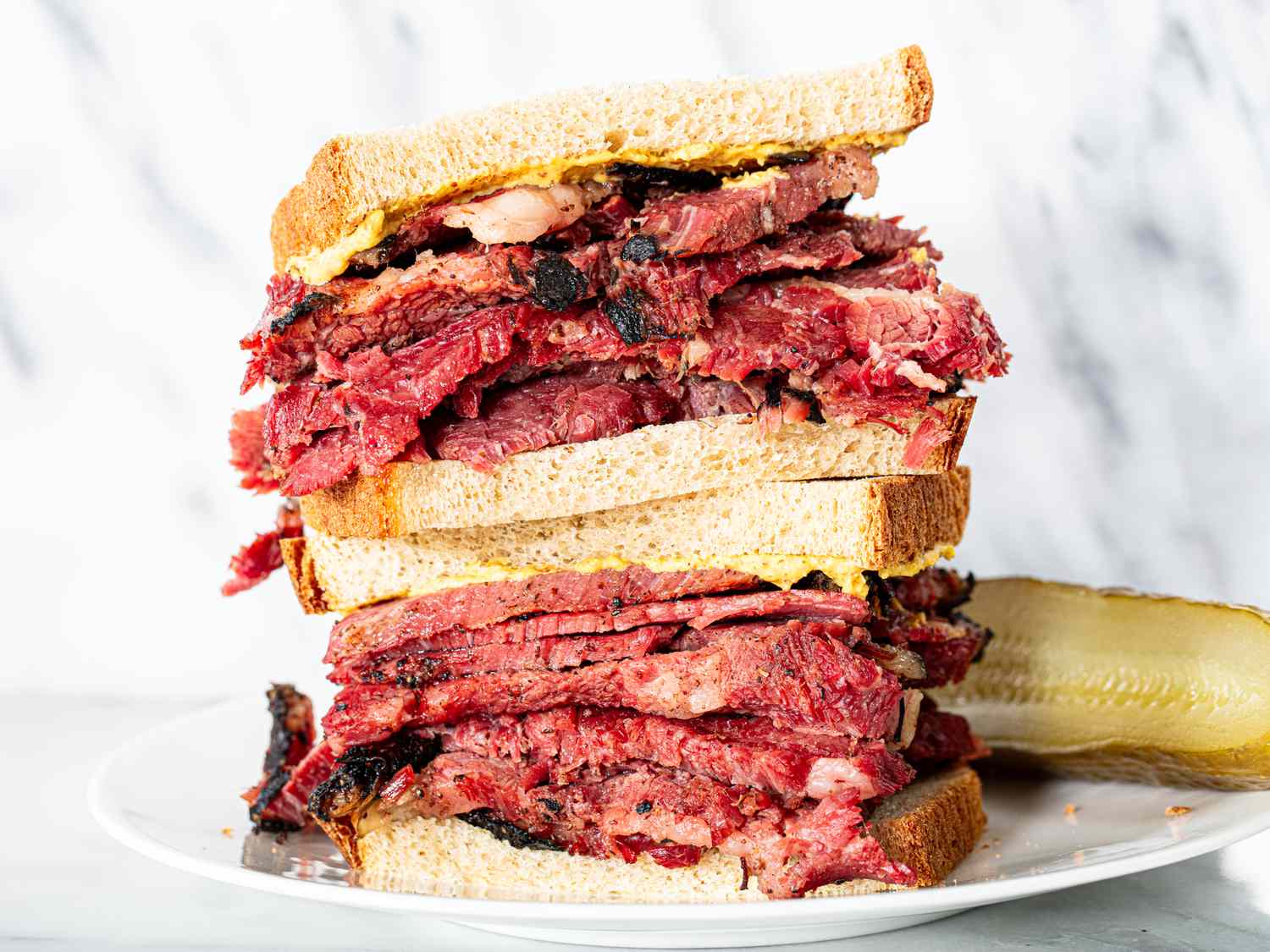 Two halves of a hulking pastrami on rye sandwich stacked on top of each other with a full sour pickle next to them on the plate.