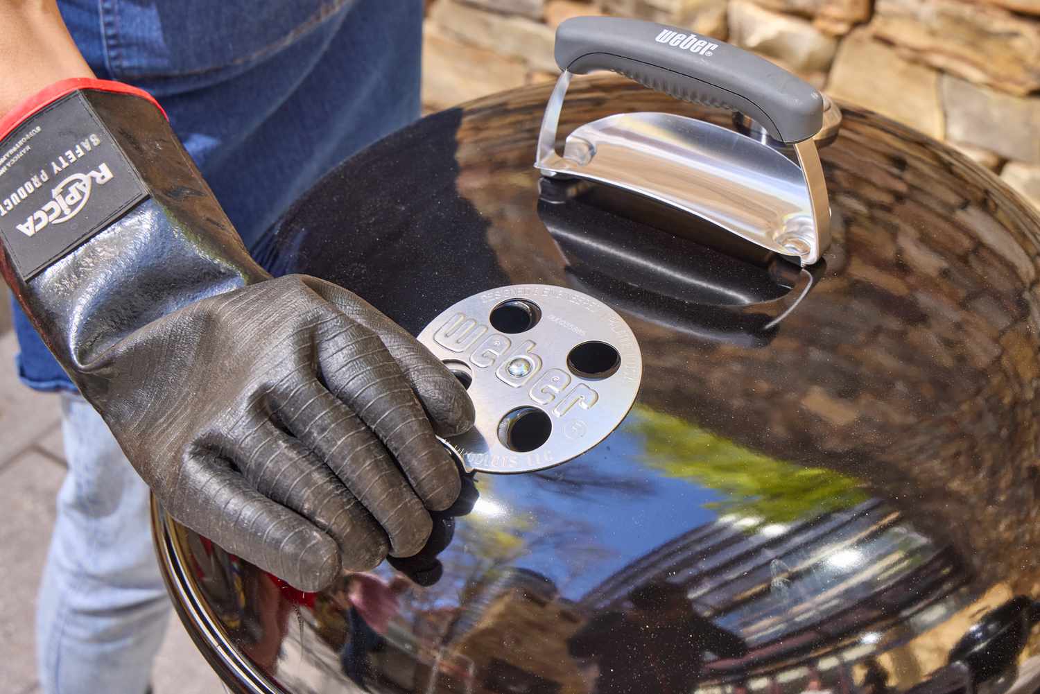 A gloved hand touching the top of the Weber Original Kettle Premium 22-Inch Charcoal Grill