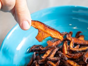A person holds a piece of cooked mushroom over a blue bowl containing more cooked mushroom pieces