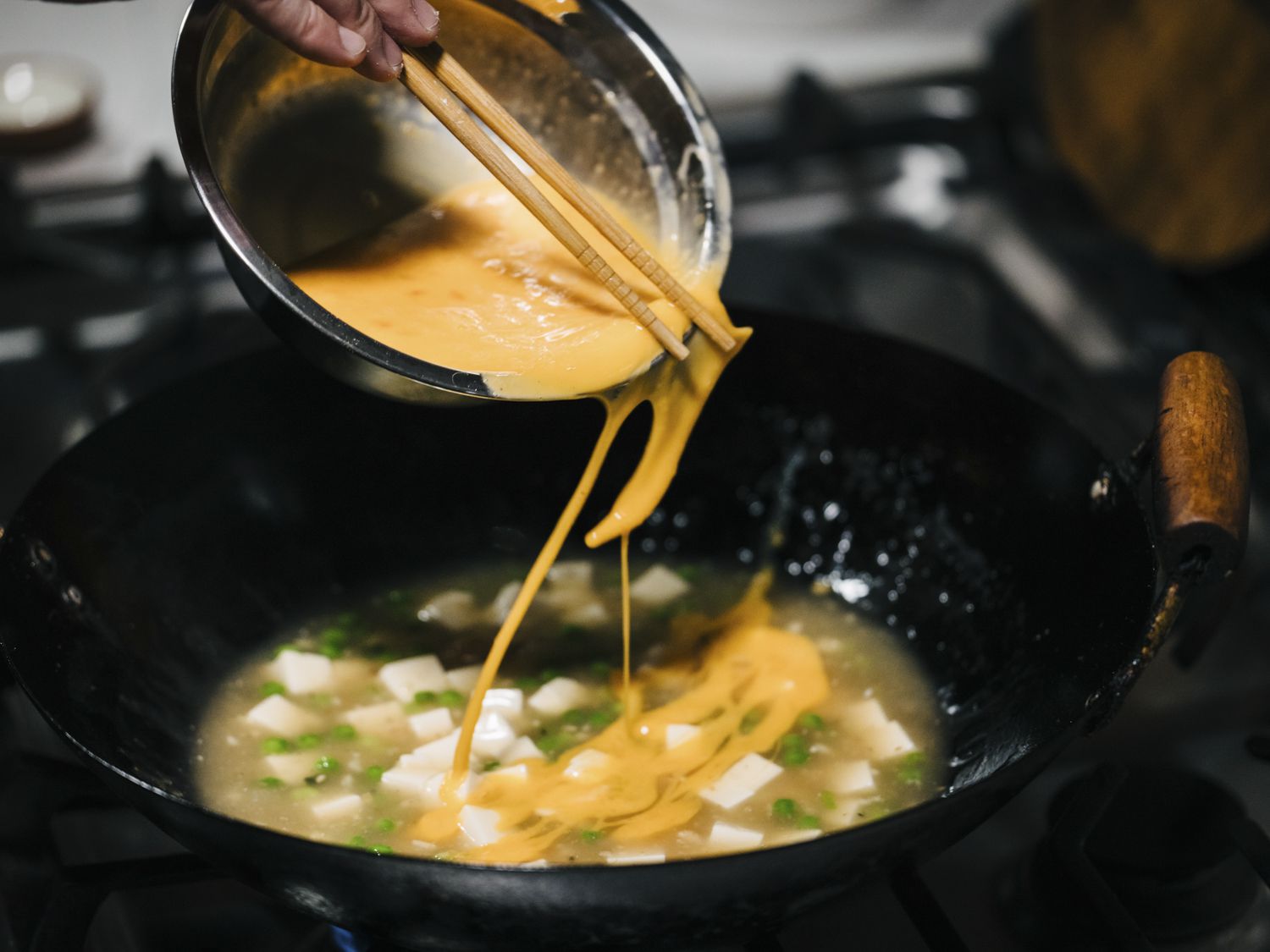 Eggs being drizzled in a swaying motion from a metallic bowl into the tofu mixture in a wok