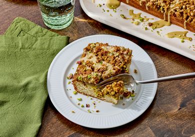 A slice of pistachio coffee cake on a plate with a fork a green napkin and glass of water nearby tray with more cake in the background