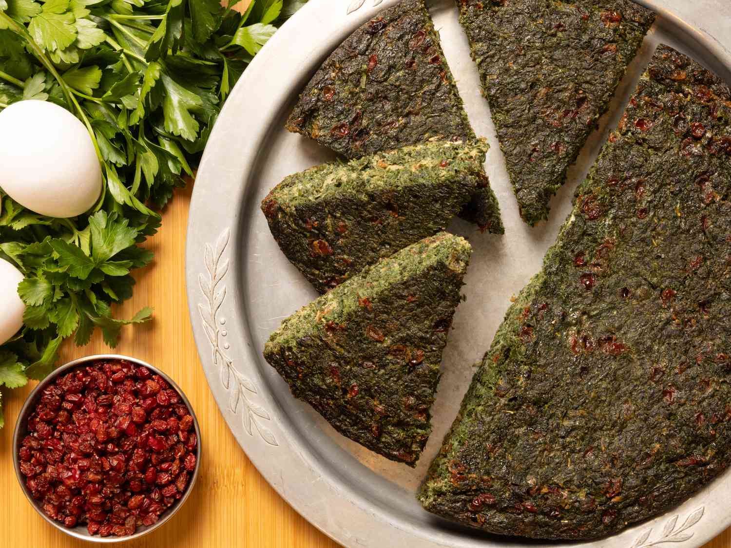 Silver platter of herb kookoo with herbs, eggs and small bowl of barberries on the side 