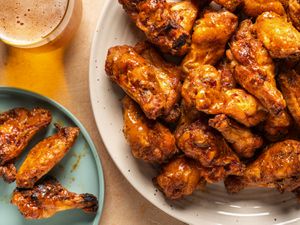 A large plate of grilled cajun chicken wings on the right hand side of the image. On the left side is a smaller blue plate with three chicken wings on it and a glass holding a glass of a golden-colored beverage.
