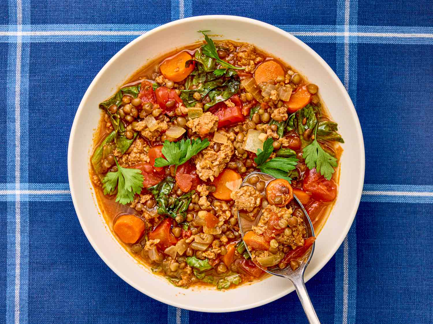 A bowl of sausage and lentil soup with vegetables garnished with parsley on a blue plaid tablecloth