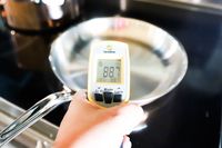 A hand holding a digital thermometer showing 887F in front of a metal frying pan
