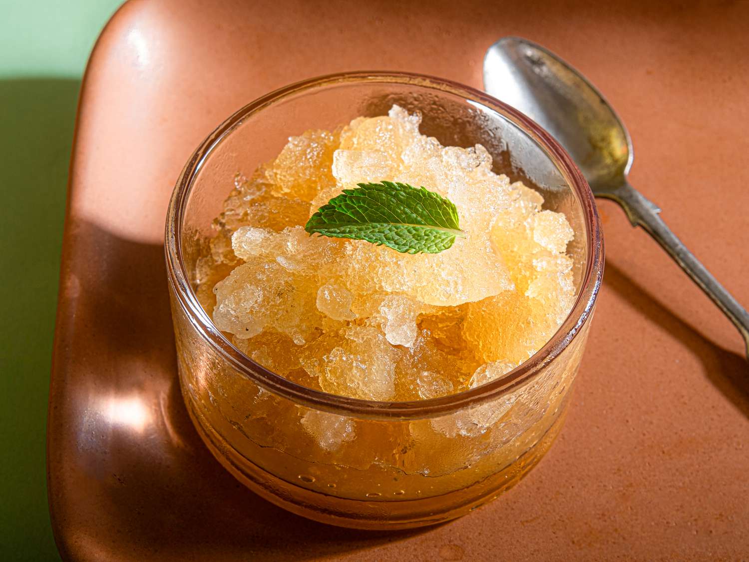 A glass bowl of granita topped with a single mint leaf, placed next to a spoon
