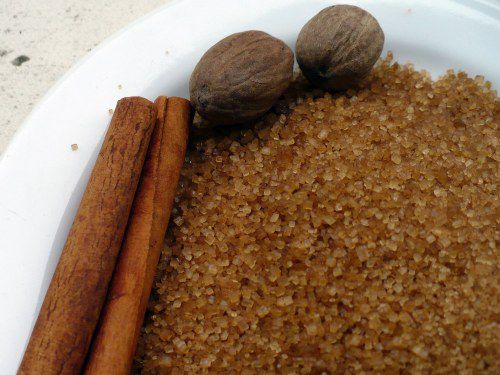 Close-up of a bowl filled with turbinado sugar, with two cinnamon sticks and whole nutmegs. 