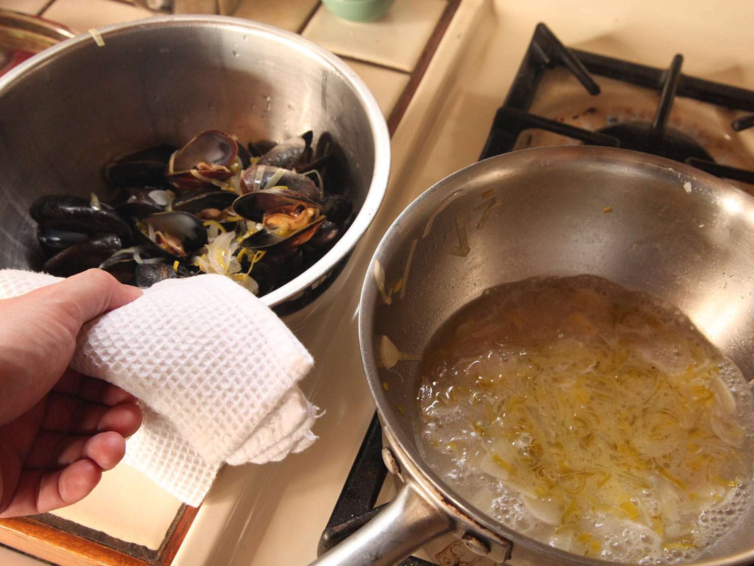 The steamed, open mussels are set aside in a bowl. The pan of mussel cooking liquid is still simmering.