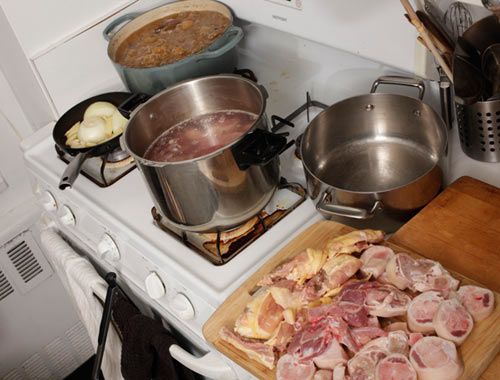 Two pots of broth and one cast iron skillet of onions cooking on a stove, a cutting board full of pork and chicken parts in the foreground.