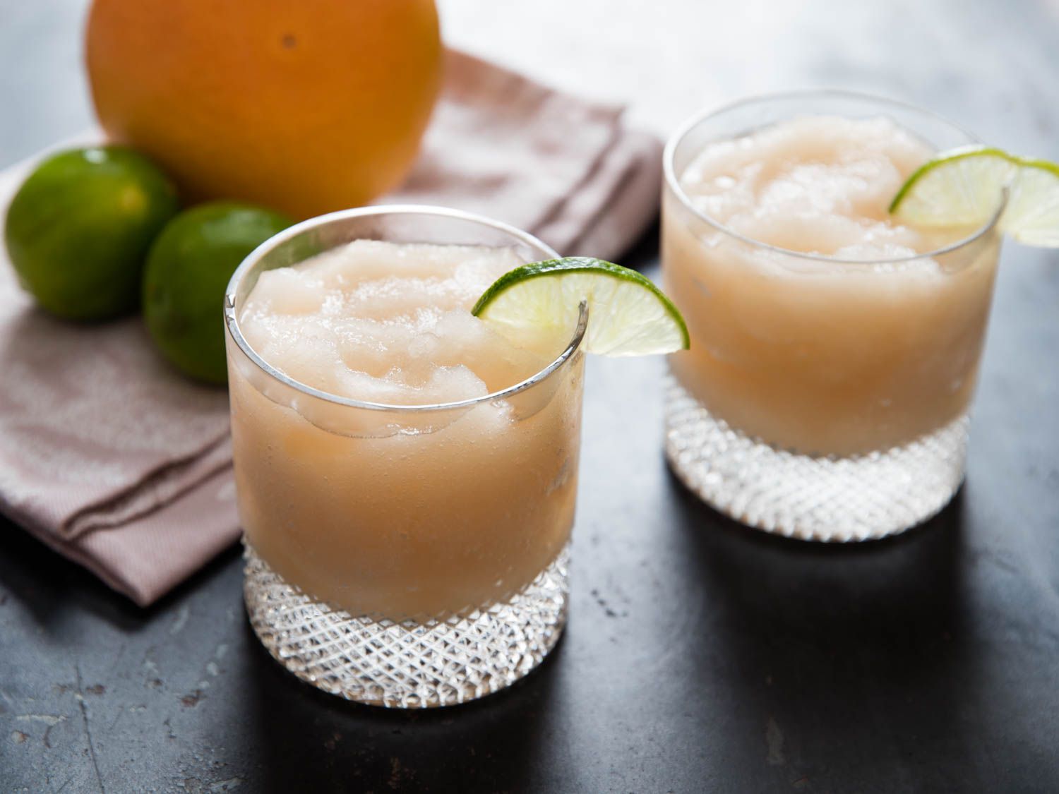 Two frozen palomas, garnished with lime slices, next to a whole grapefruit and two whole limes.