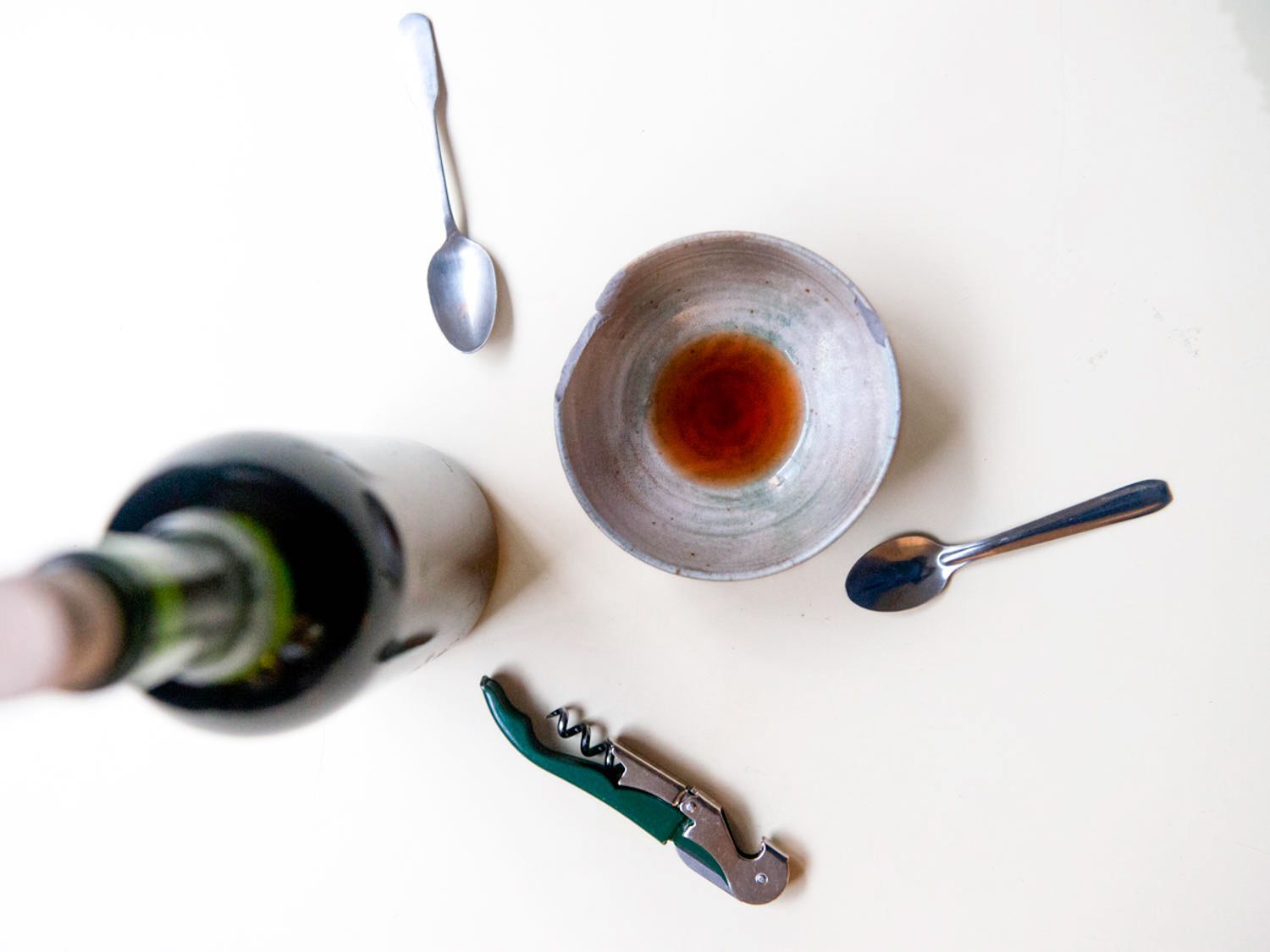 Overhead view of a small earthenware bowl for sampling vinegar. An uncorked bottle of vinegar is nearby, as are two tasting spoons.