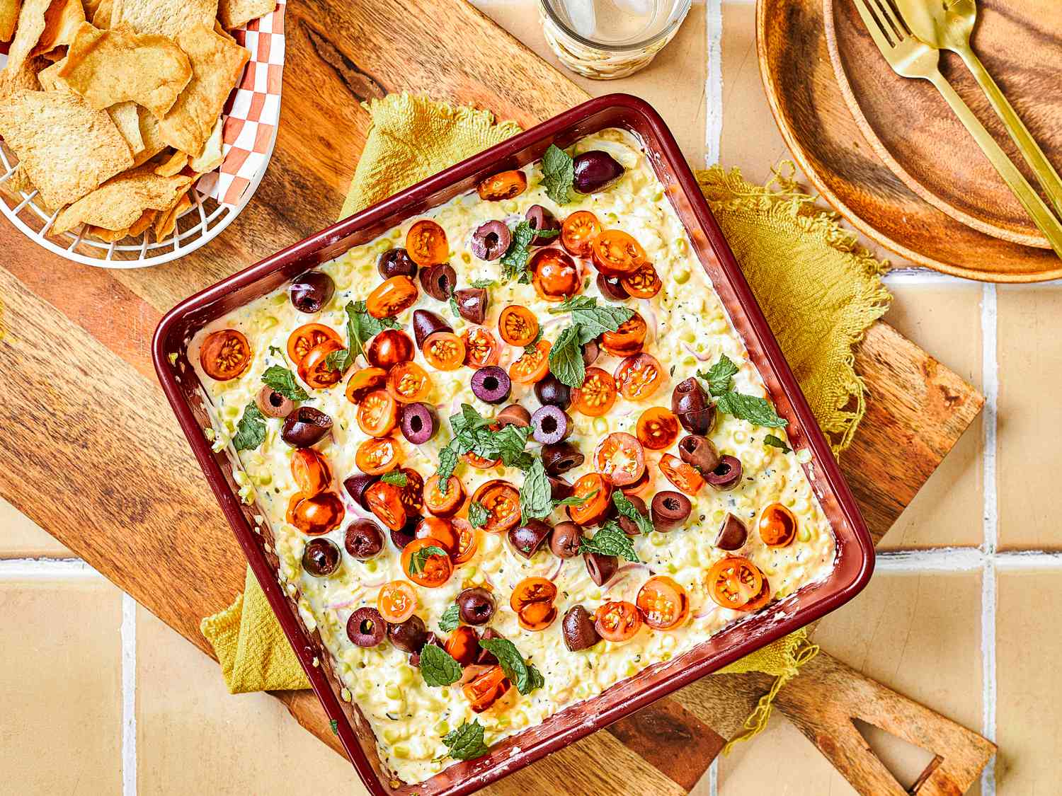 Overhead view of corn dip topped with olives, mint, and cherry tomatoes on a table with chips, a drink, and a wooden board.