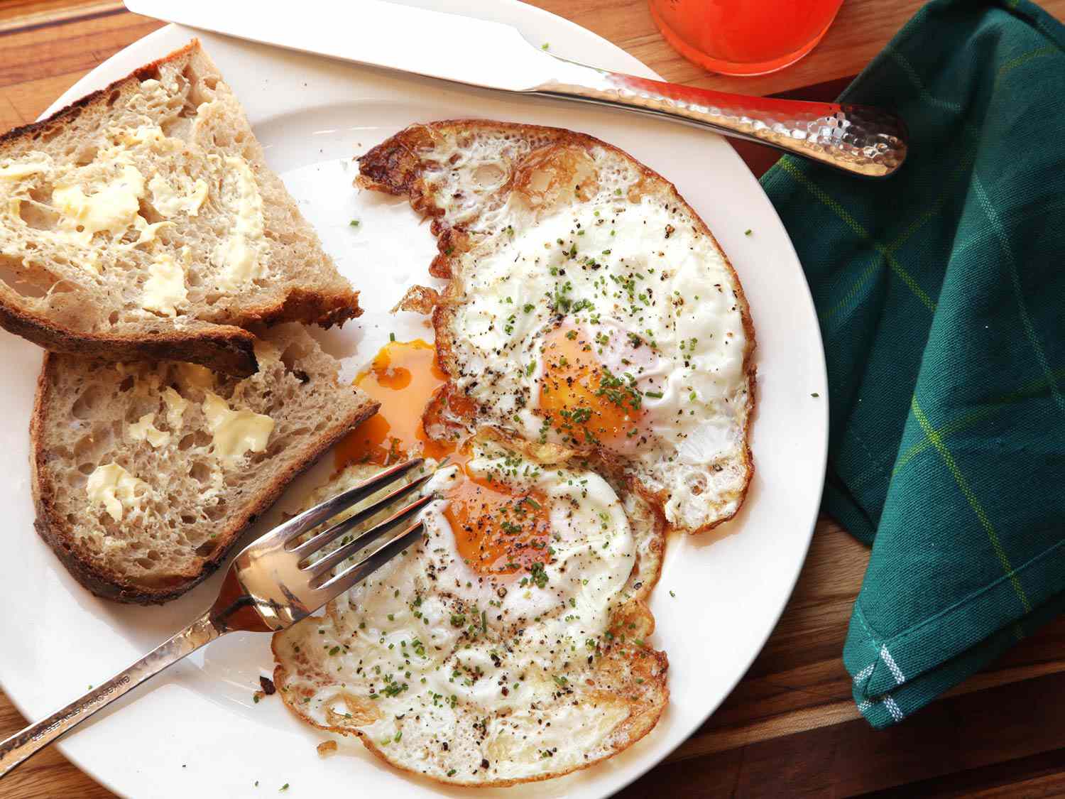 Two crisp fried eggs plated with slices of buttered toast; one yolk has been broken and is oozing. 
