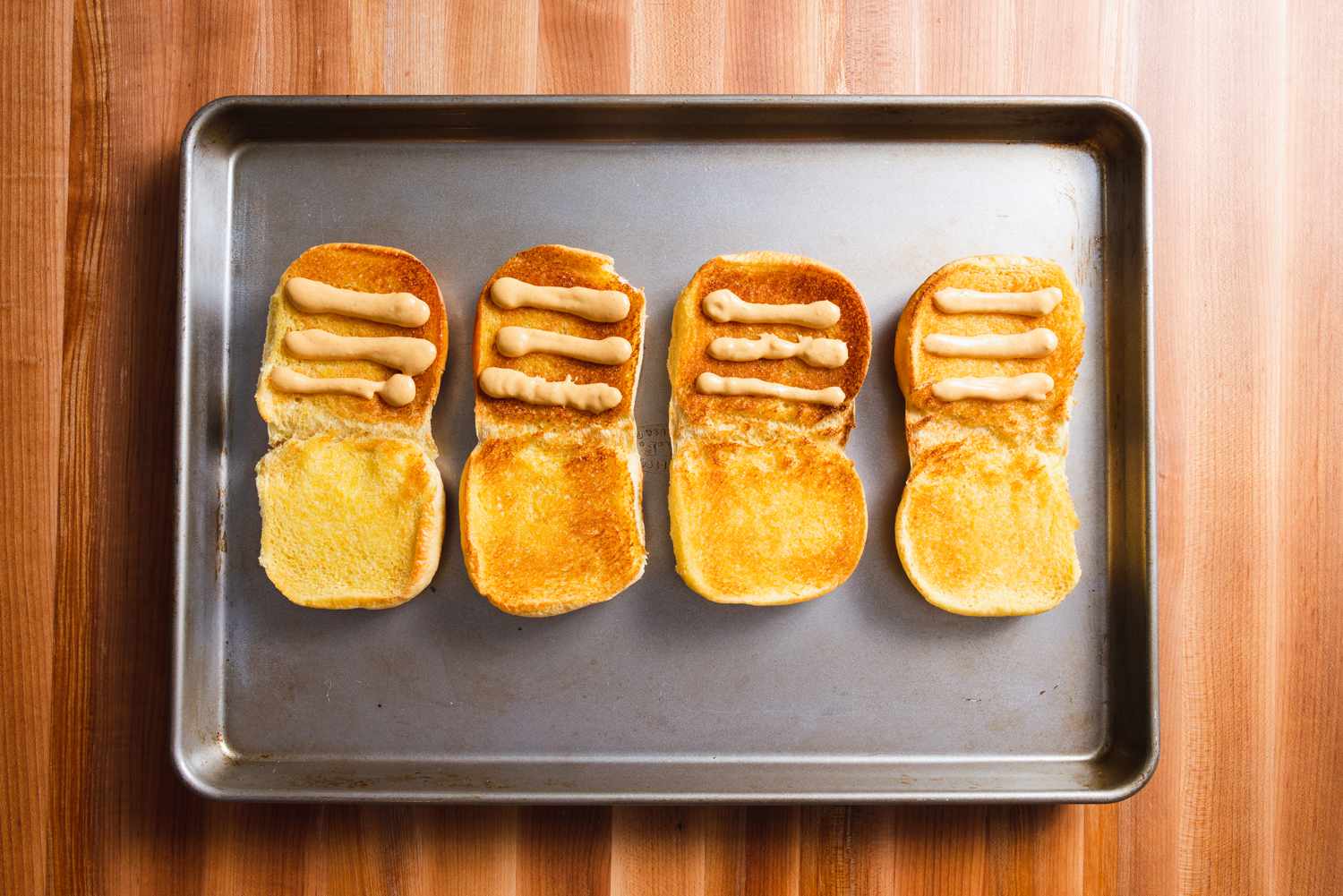 Four toasted burger buns open on a baking sheet, each one with three lines of homemade Shack Sauce on them.