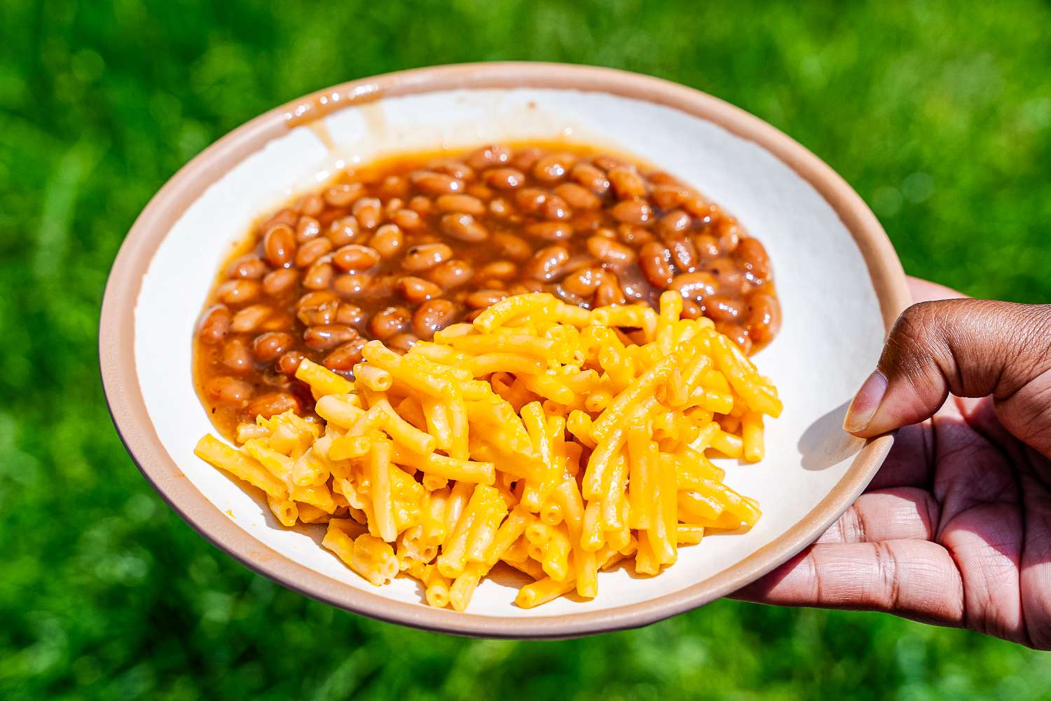 A hand holding a Lulu & Georgia Tara Melamine Dinnerware Set plate with baked beans and macaroni and cheese on a grassy background