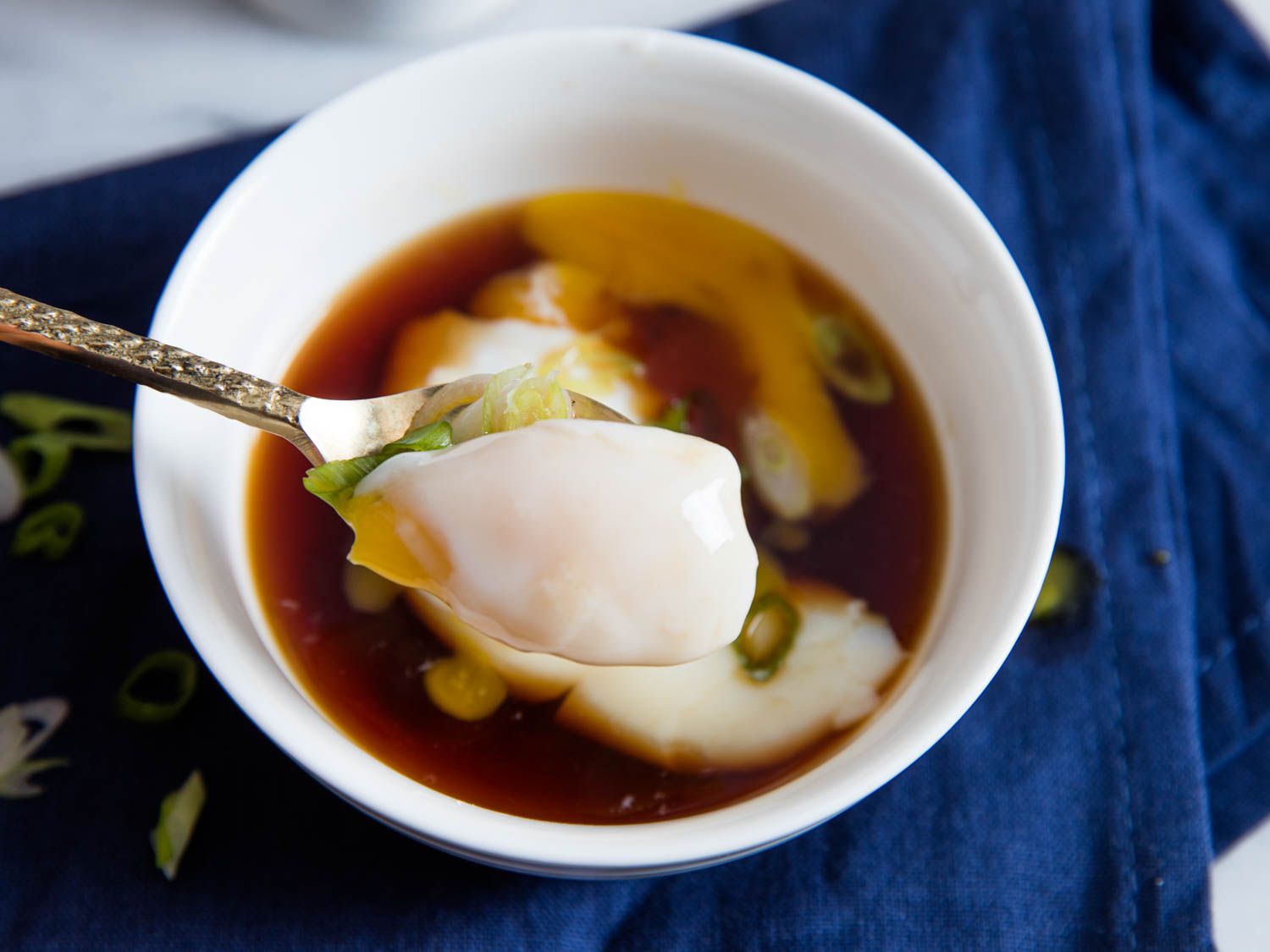 A spoon holding a bight of the onsen egg close to the camera. The serving bowl is in the frame below the bite, blurred by the depth of focus. Egg yolk has pooled into the broth.