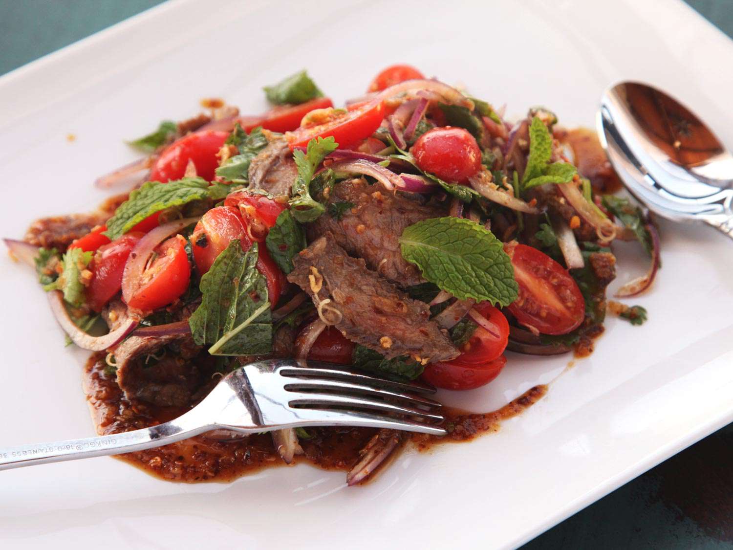 Isan Thai–style sliced-steak salad with herbs, tomato, and onion, on a white plate with a fork