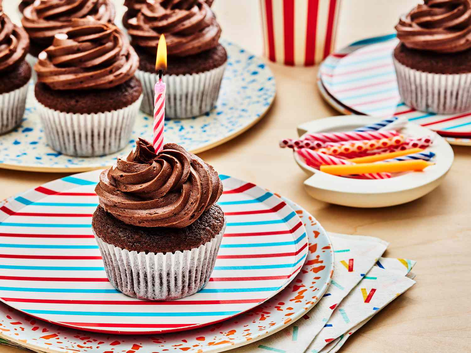 Chocolate cupcake with lit candle in the frosting on a stack of colorful plates. In the background, there is a plate of more cupcakes and a bowl of birthday candles. 
