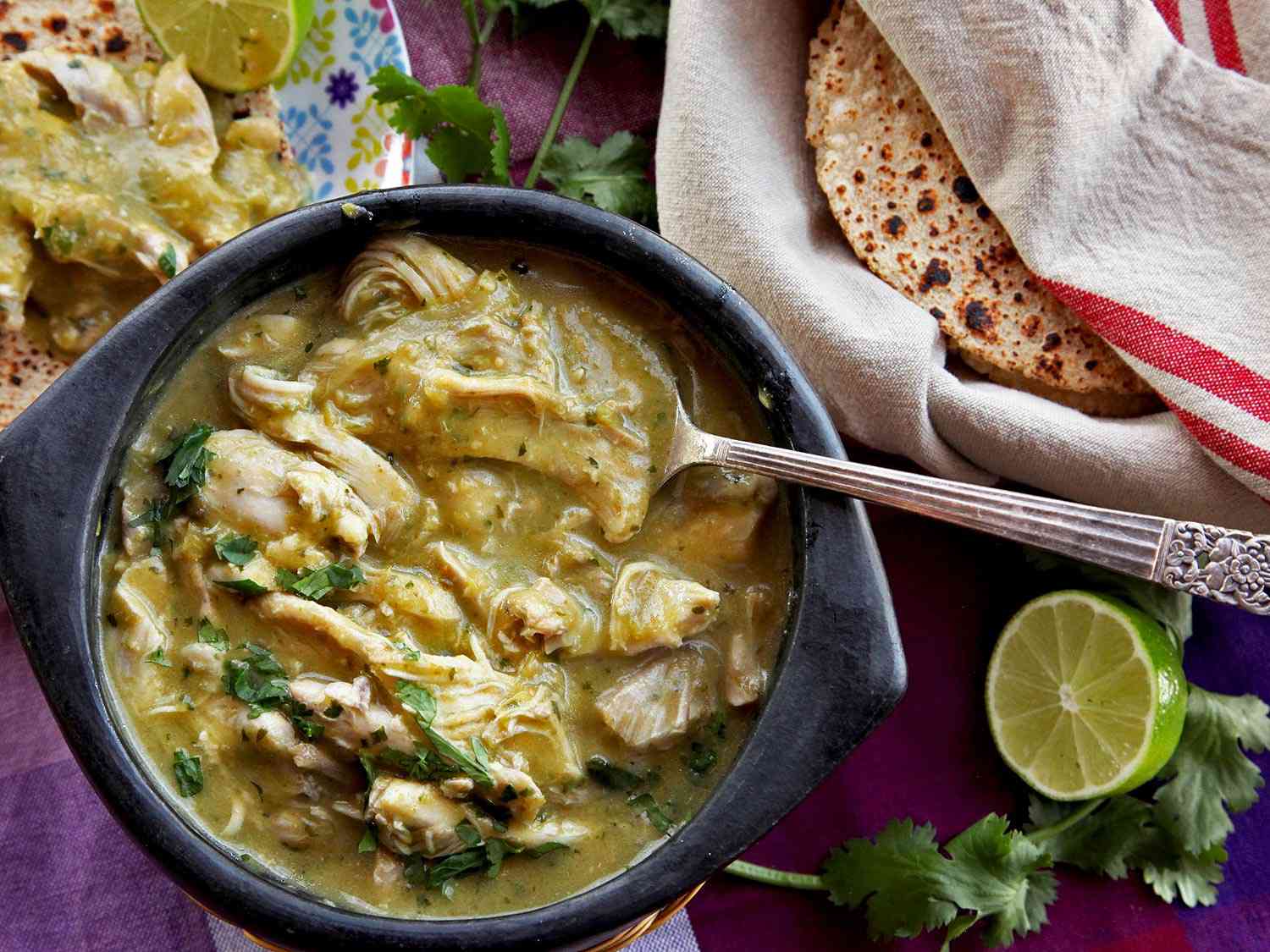 Overhead view of a bowl of chicken chile verde, served in a black earthenware bowl with corn tortillas, lime halves, and cilantro.