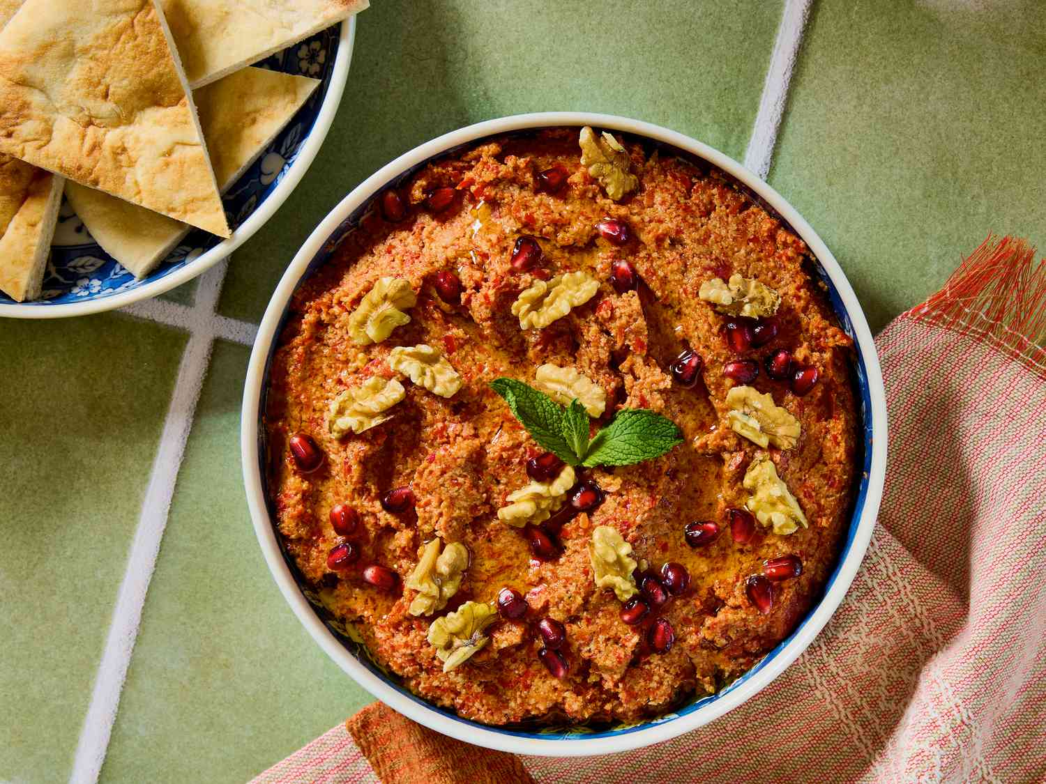 A bowl of muhammara garnished with walnuts, pomegranate arils, and a mint leaf, next to pita bread on a green tile surface