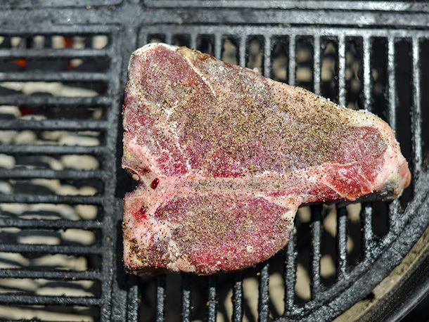 Overhead shot of a seasoned, raw porterhouse that's been placed on the cooler side of the grill, positioned so that the T-bone side is closest to the coals.