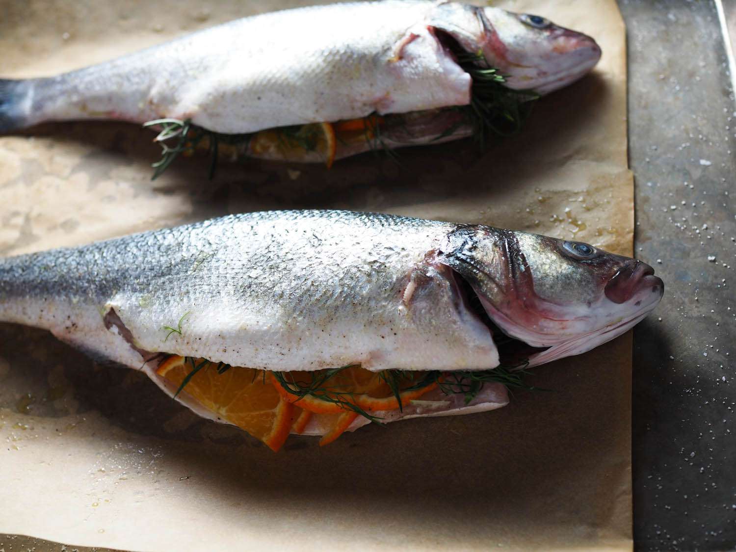 Two whole branzini, bellies stuffed with fennel fronds, rosemary, and tangerine slices, laying on brown parchment paper.