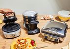 Three waffle makers on a counter, surrounded by breakfast items