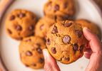 A closeup of hands holding a chocolate chip cookie with other cookies visible on a plate in the background
