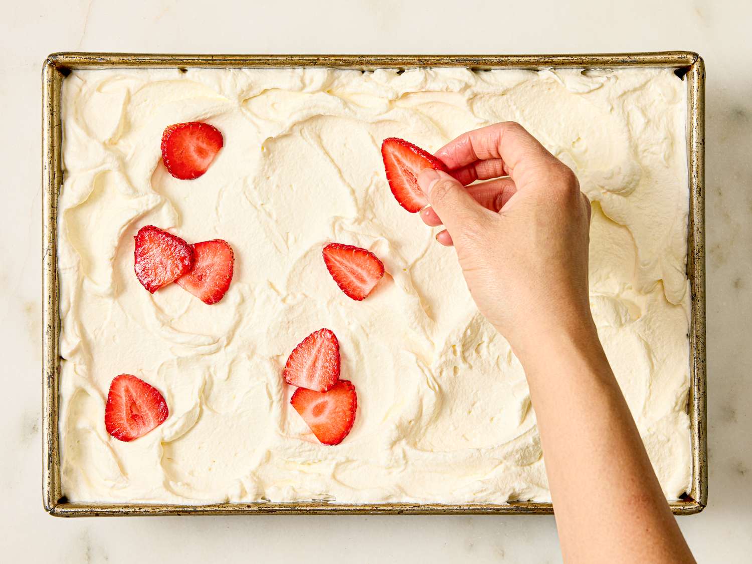 Hand placing strawberry slices on a dessert with a whipped topping layer in a baking dish