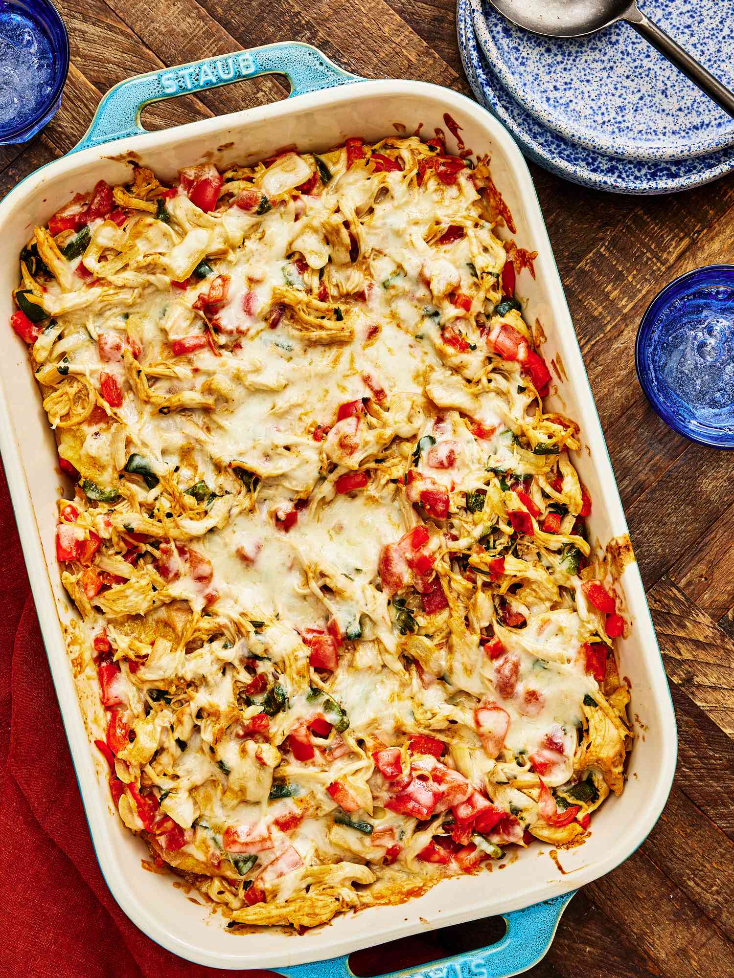 Large blue baking pan of whole King Casserole on a wooden surface, with blue splatter dishes, red napkins, and glasses of sparkling water