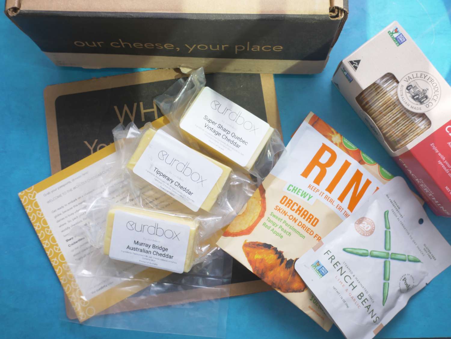 Three small blocks of cheese, a box of crackers, and bags of dried fruit and French beans on a blue surface