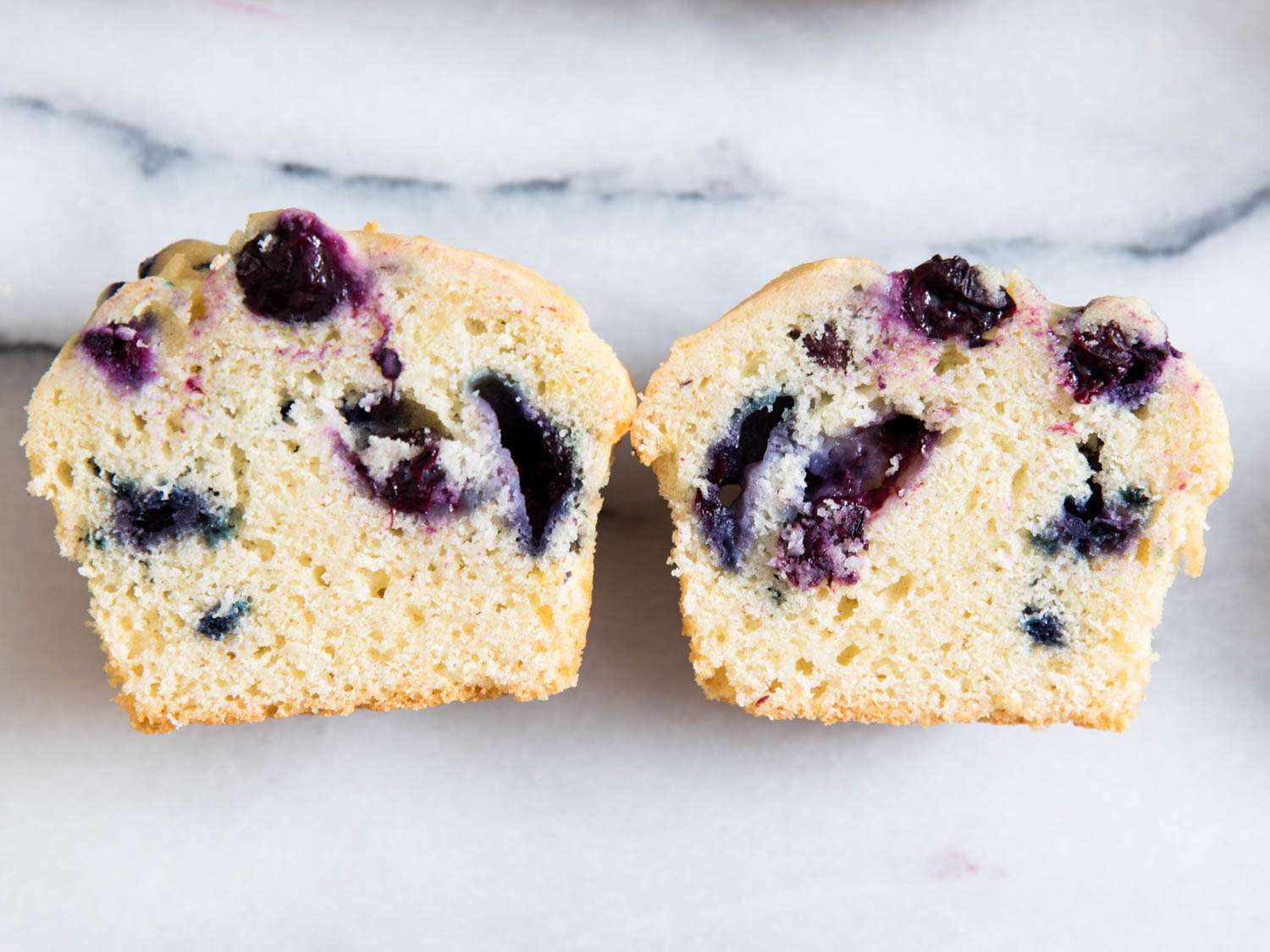 A close up of a blueberry muffin cut in half, with bursting berries evenly distributed.