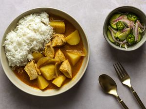 A bowl of Thai chicken yellow curry in a cream colored ceramic bowl. On the right side of the image is a smaller bowl holding the accompanying cucumber salad. There are a fork and spoon in the bottom right hand corner of the image.