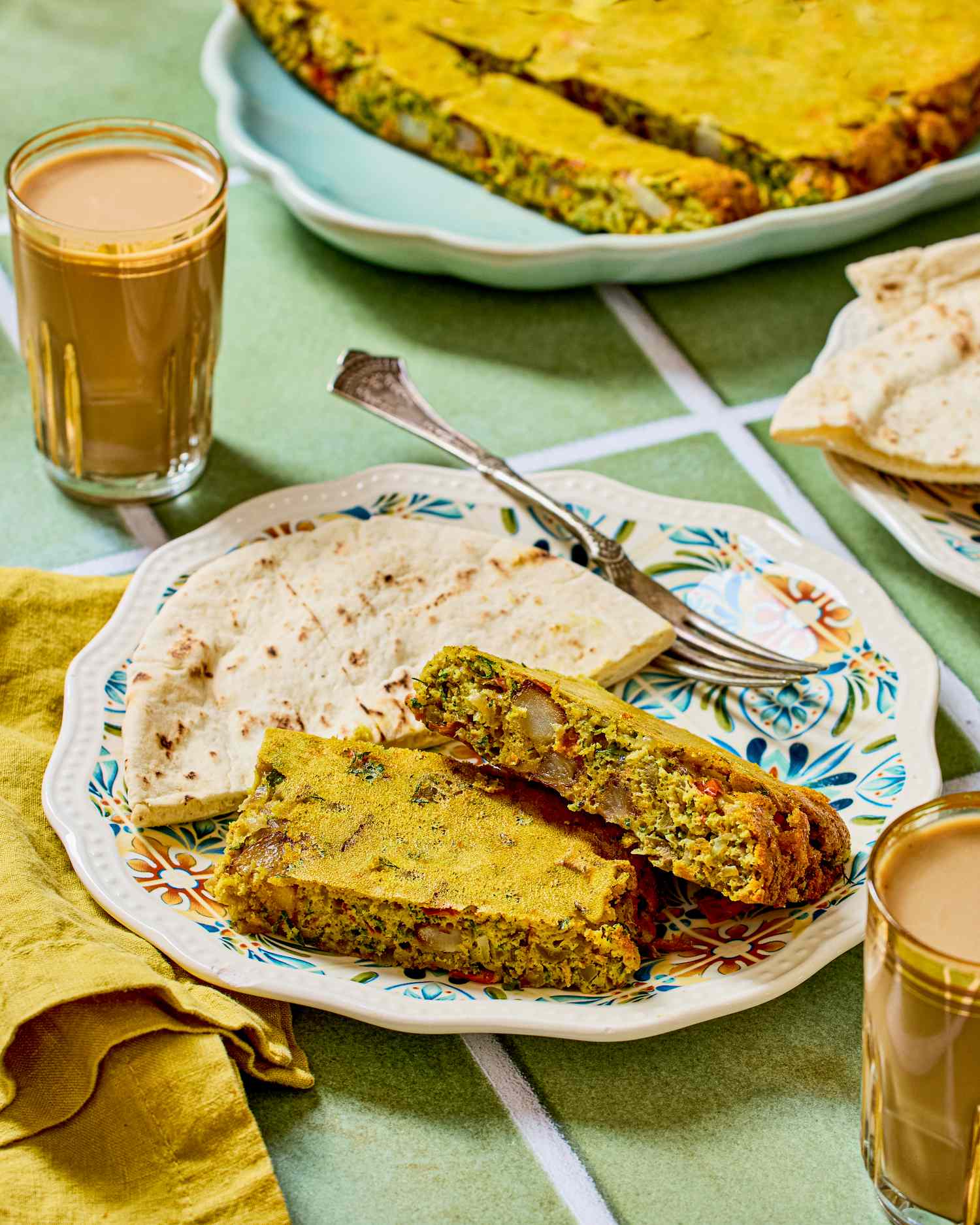 Slices of savory baked dish on a patterned plate with flatbread and glasses of tea on the side
