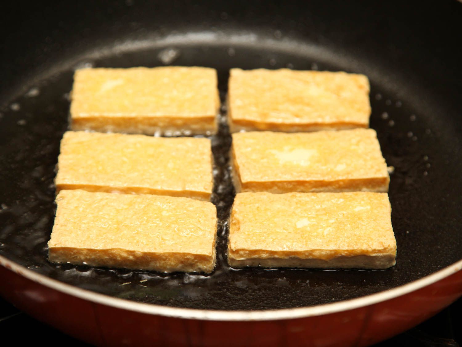 Six tofu slices frying in a skillet, browned on top side.
