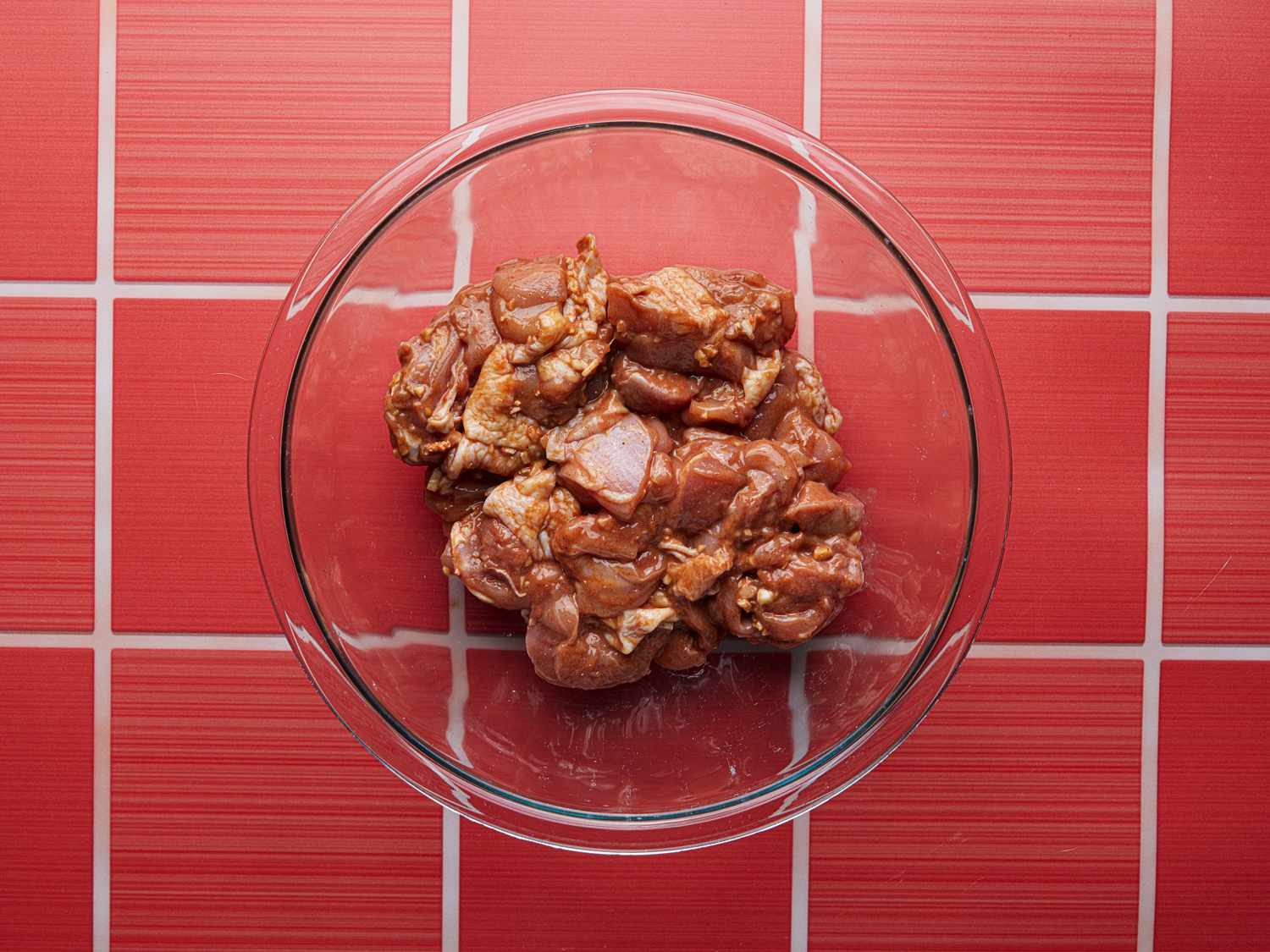Overhead view of marinaded chicken in a glass bowl on a red counter