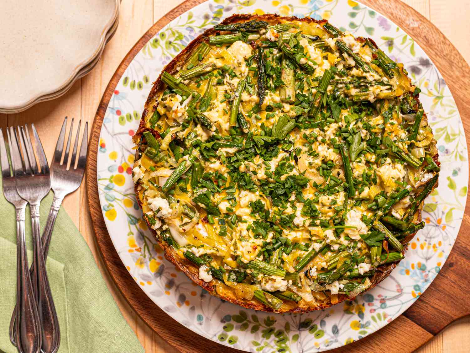 Smashed potato tart with asparagus and herbs on a floral plate, placed on a wooden table with forks and a green napkin nearby.