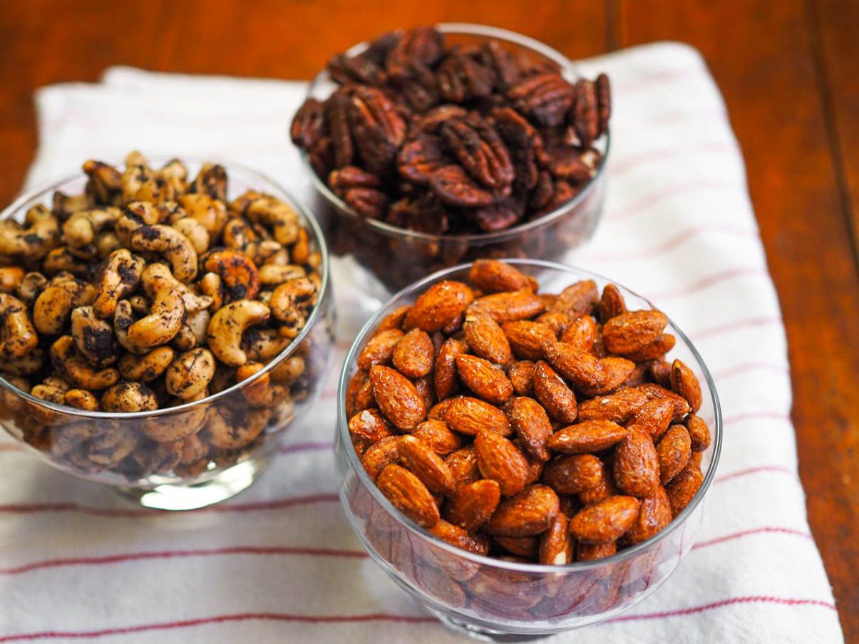 Three glass dishes of spiced nuts on a kitchen towel on a table. The flavors are smoky candied almonds, Mexican spiced chocolate pecans, and olive-rosemary cashews.