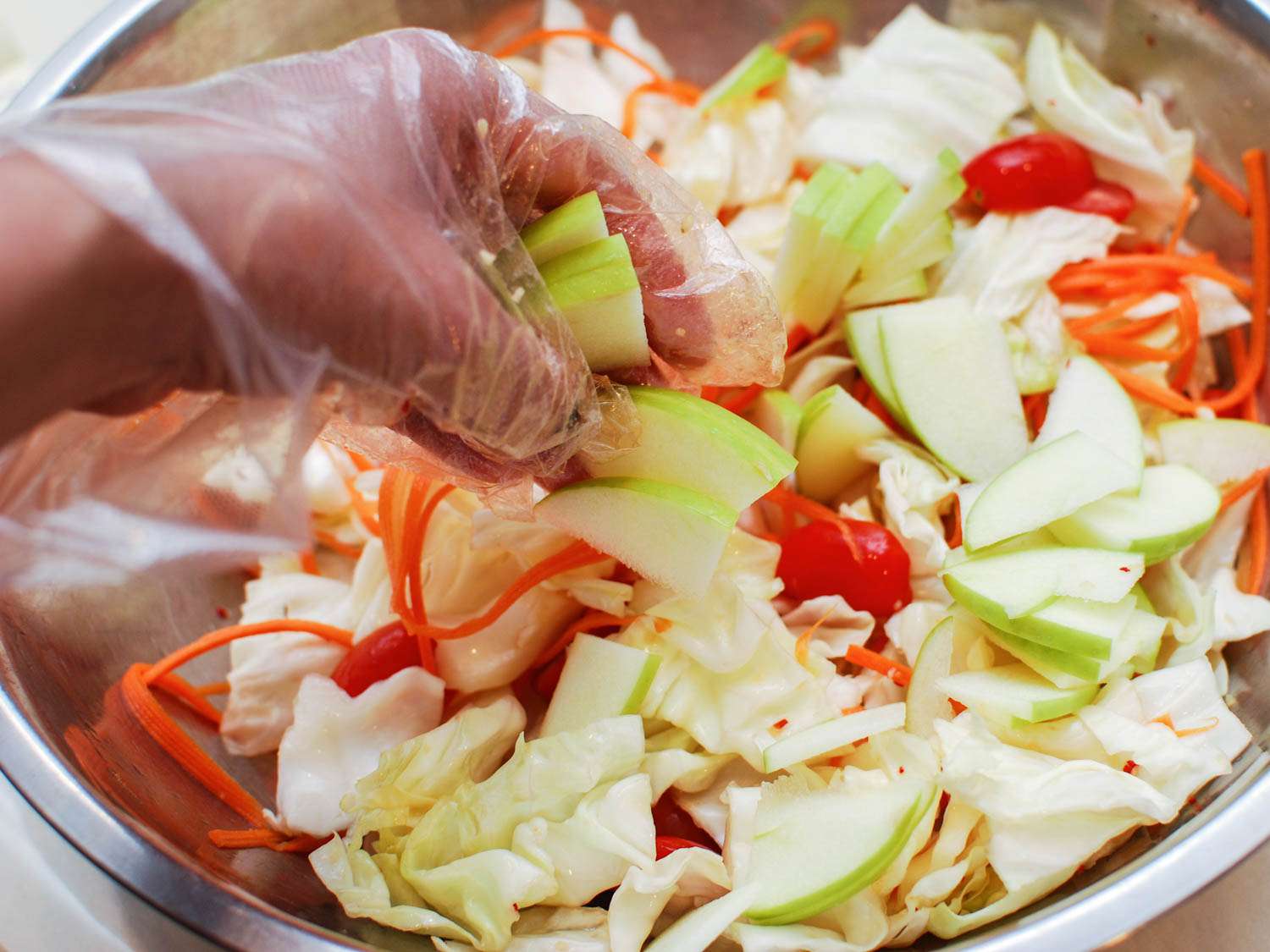 Adding sliced apple to a salad bowl with cabbage, julienned carrot, and cherry tomatoes.