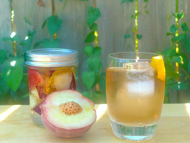 A canning jar of DIY peach liqueur next to a stirred cocktail and a fresh, halved peach.