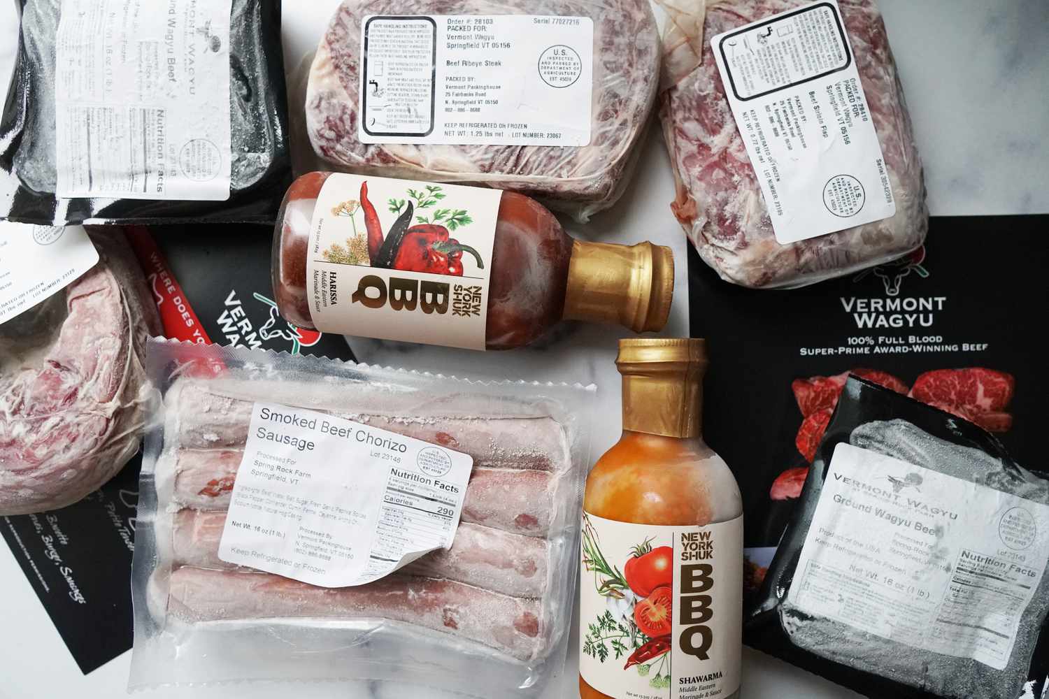 Blocks of frozen meat, a package of ground beef, a package of chorizo links, and two bottles of barbecue sauce on a marble surface